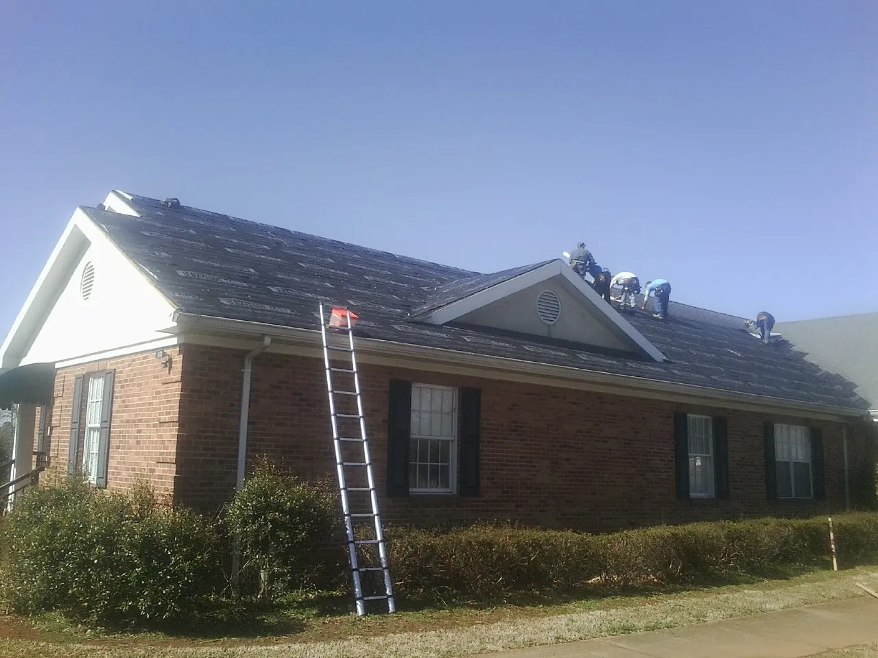 Crew tearing shingles off a brick ranch roof with a ladder set — Metal Roof Repair work by our crew in Riesel